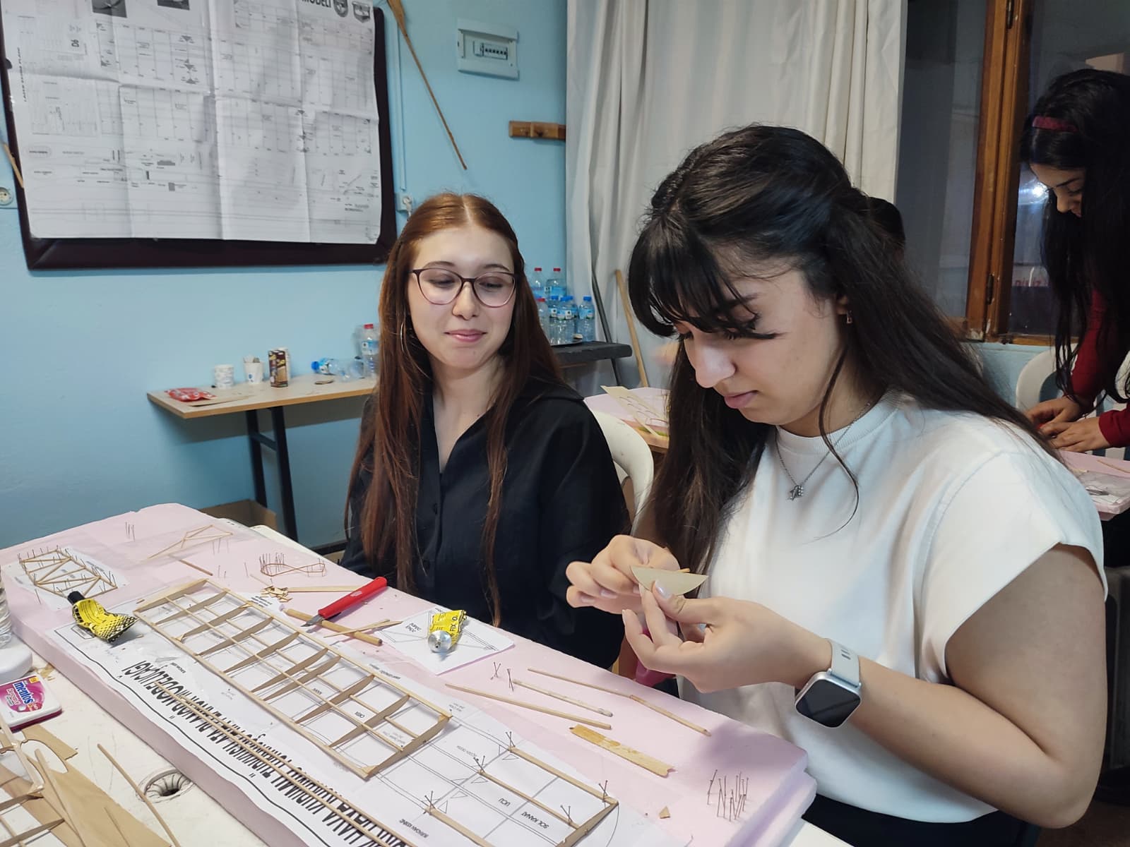 Öğrencilerin atölyede model uçak yapımıyla ilgilendiklerini gösteriyor. Öğrenciler, eğitmenlerin yardımıyla uçak parçalarını bir araya getiriyor, yapıştırıyor ve şekillendiriyor. Atölye ortamı, öğrencilerin öğrenmeye istekli ve keyifli bir şekilde çalıştıklarını yansıtıyor.